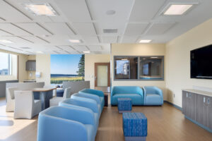 Modern healthcare patient lounge with blue lounge seating, acoustic ceiling panels, nature mural, and integrated technology reflecting current healthcare construction trends