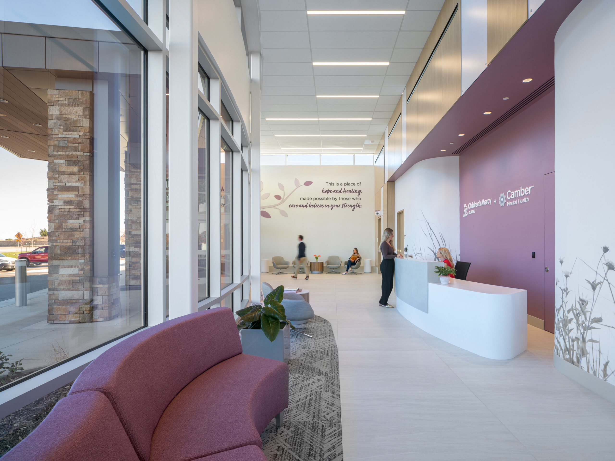 Bright hospital lobby with a curved white reception desk, purple seating, and large glass walls with people waiting nearby.