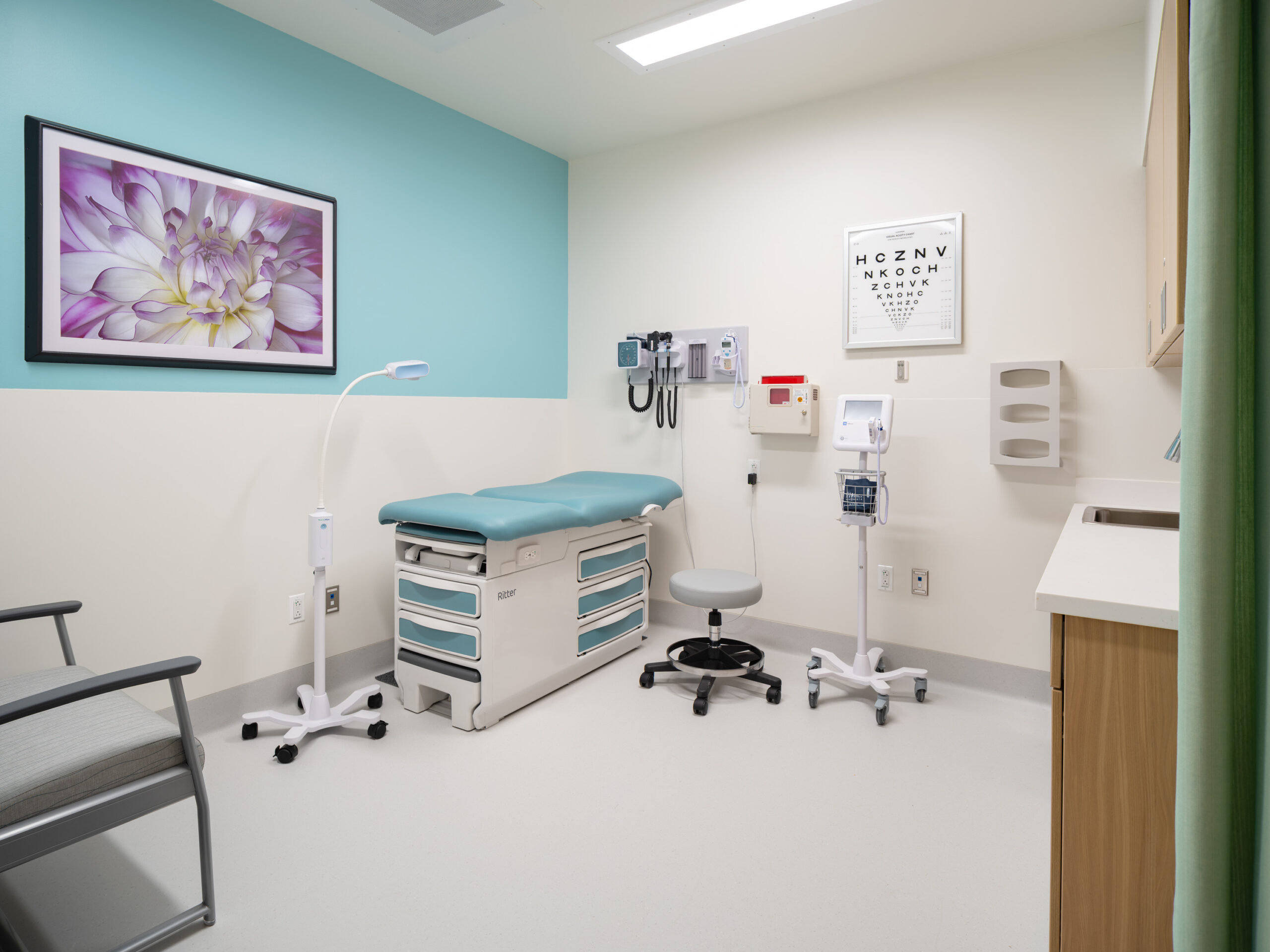 Empty medical examination room with teal wall, exam bed, and medical equipment shelf and chart on wall