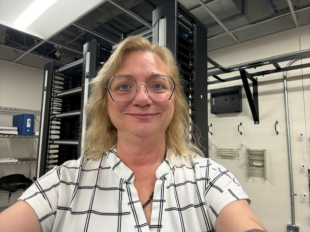 Smiling woman wearing glasses and a white checkered shirt taking a selfie in a data center with server racks in the background.