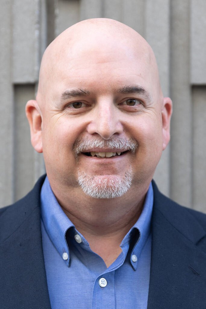Smiling bald man with gray goatee in a blue shirt and dark blazer, outdoor gray wall behind him.