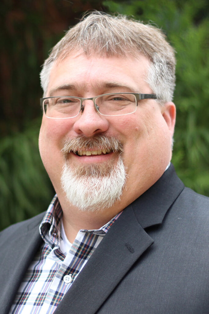Smiling man with gray hair and beard wearing glasses, a plaid shirt, and a dark blazer outdoors.