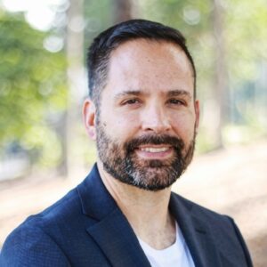 Portrait of a man with short dark hair and a beard, wearing a navy blazer, outdoors in a sunlit park.