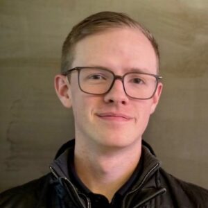 Portrait of a young man with short light hair and black-framed glasses, wearing a dark zip jacket and a subtle smile, facing the camera.