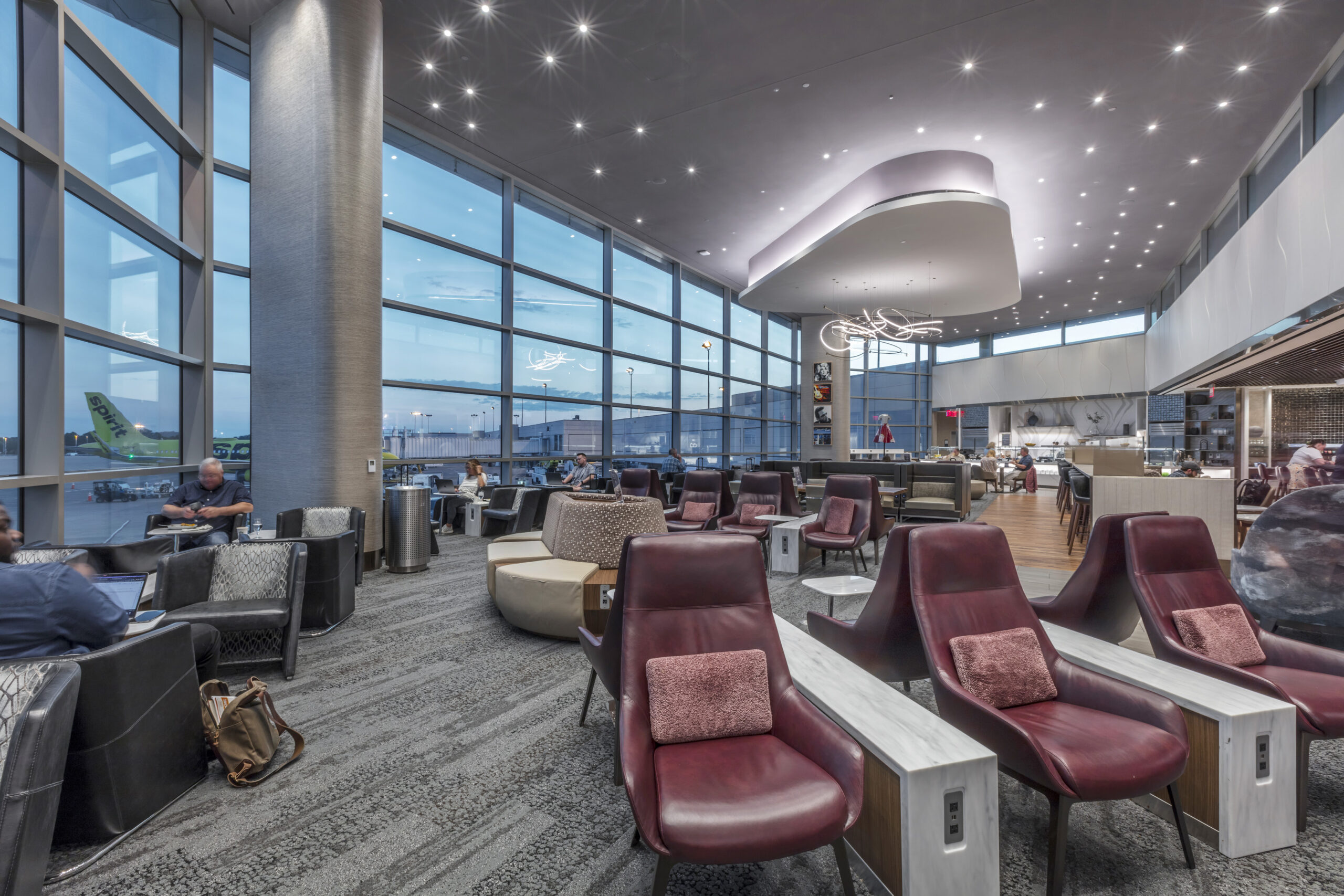 Airport lounge with red chairs, large glass windows, and a curved ceiling feature at dusk.