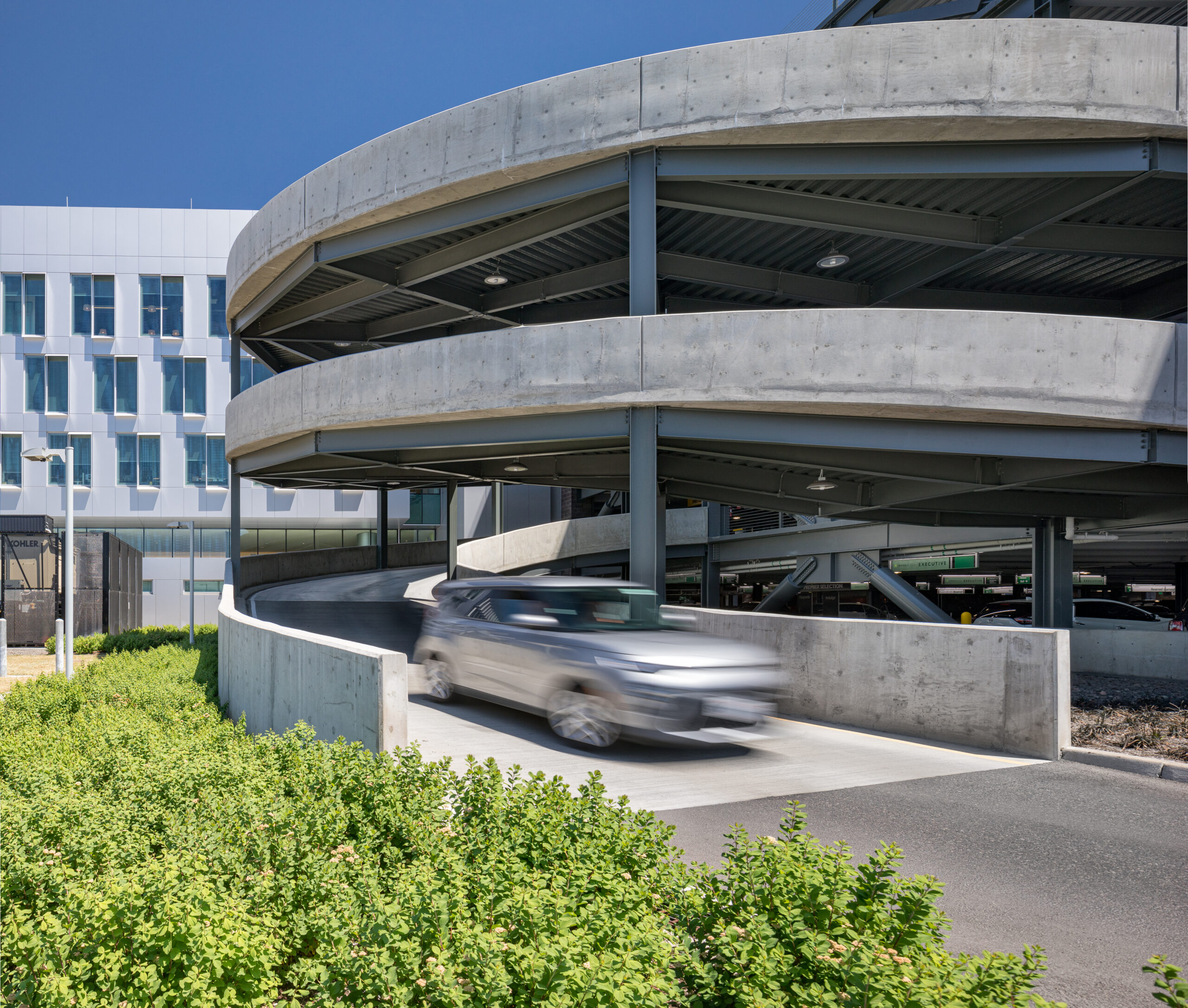 Spiral concrete parking garage with metal supports beside a white modern building under a blue sky; a blurred car passes by in the foreground.