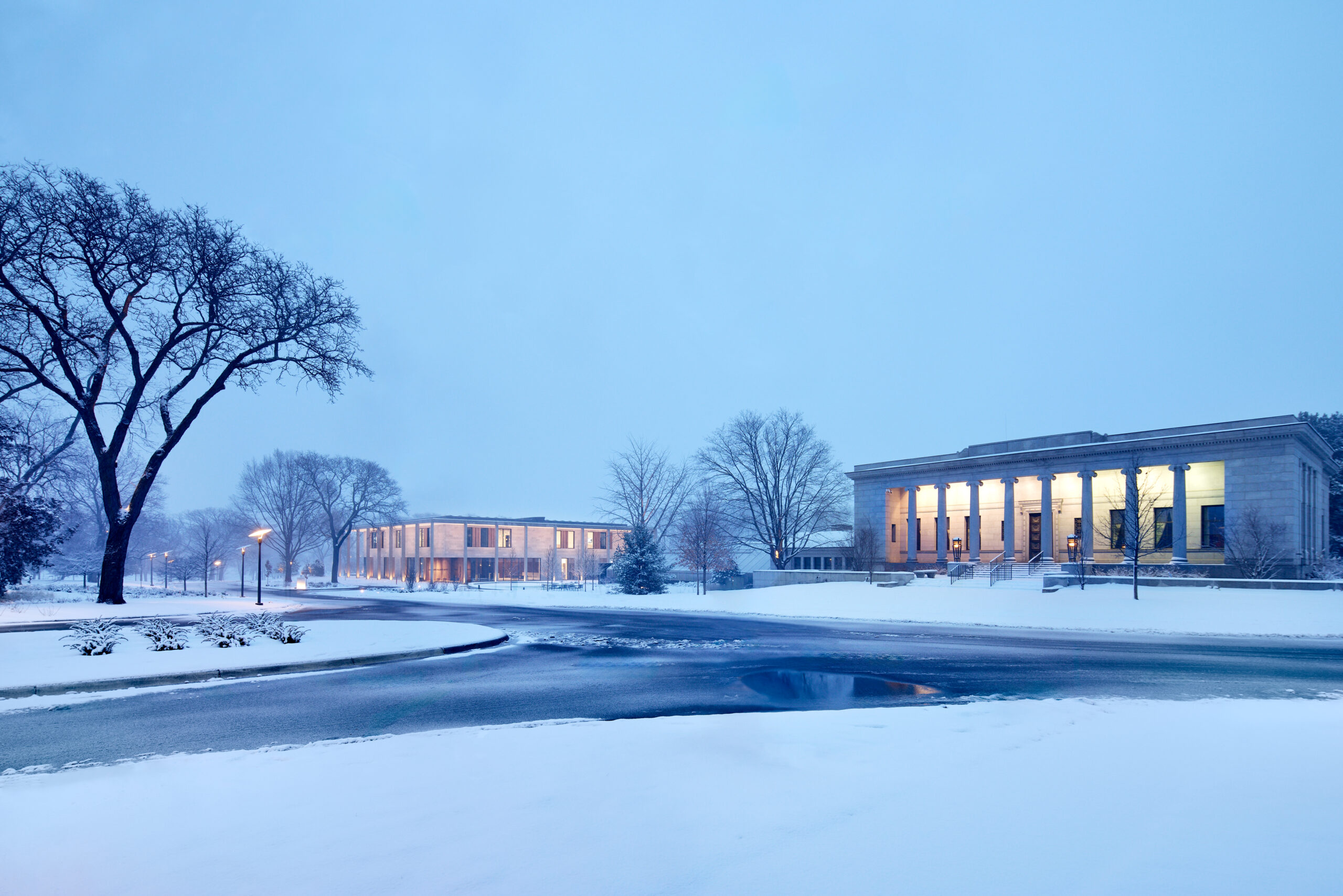 Snow-covered urban plaza at dusk with a neoclassical building on the right and bare trees in the foreground, blue twilight sky above.