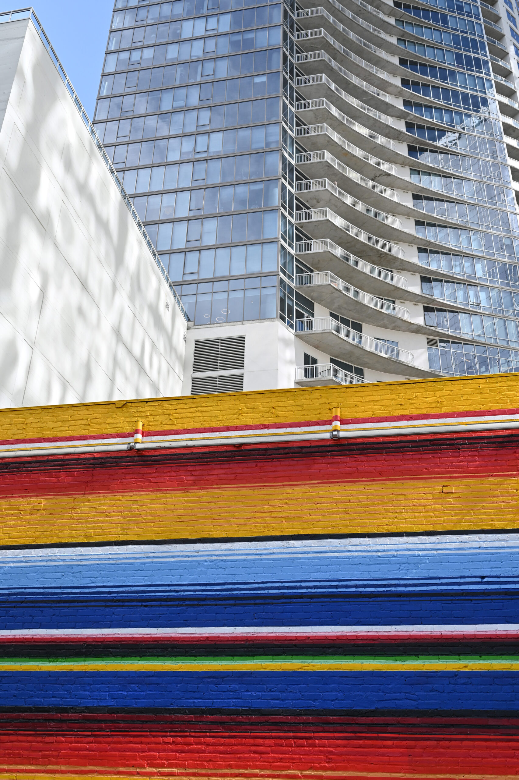Modern glass high-rise with curved balconies on the right, seen behind a vibrant striped wall in yellow, red, blue, and green in the foreground.