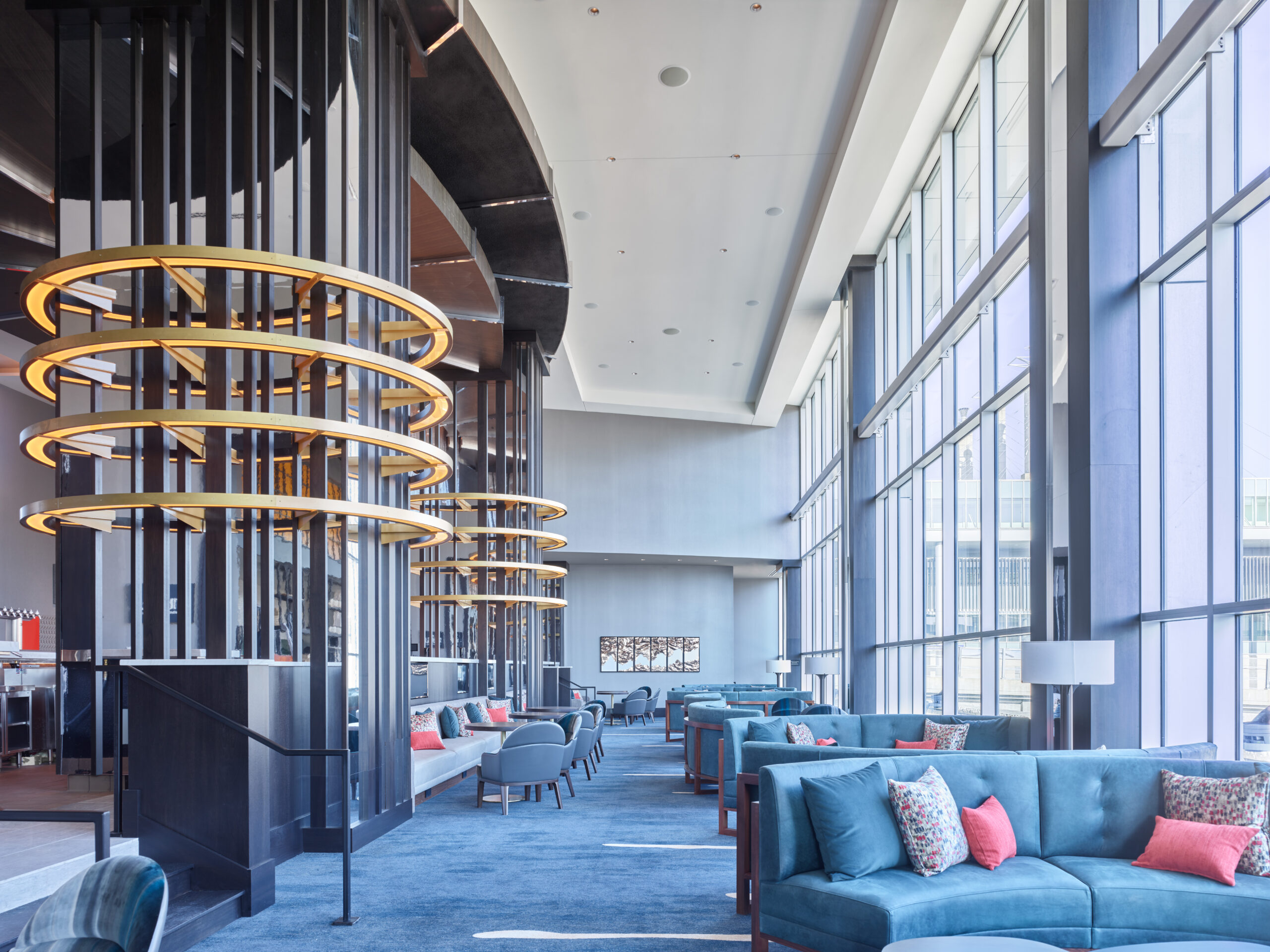 Spacious modern lobby with blue sofas and colorful cushions, circular amber light fixtures, and floor-to-ceiling windows on the right.