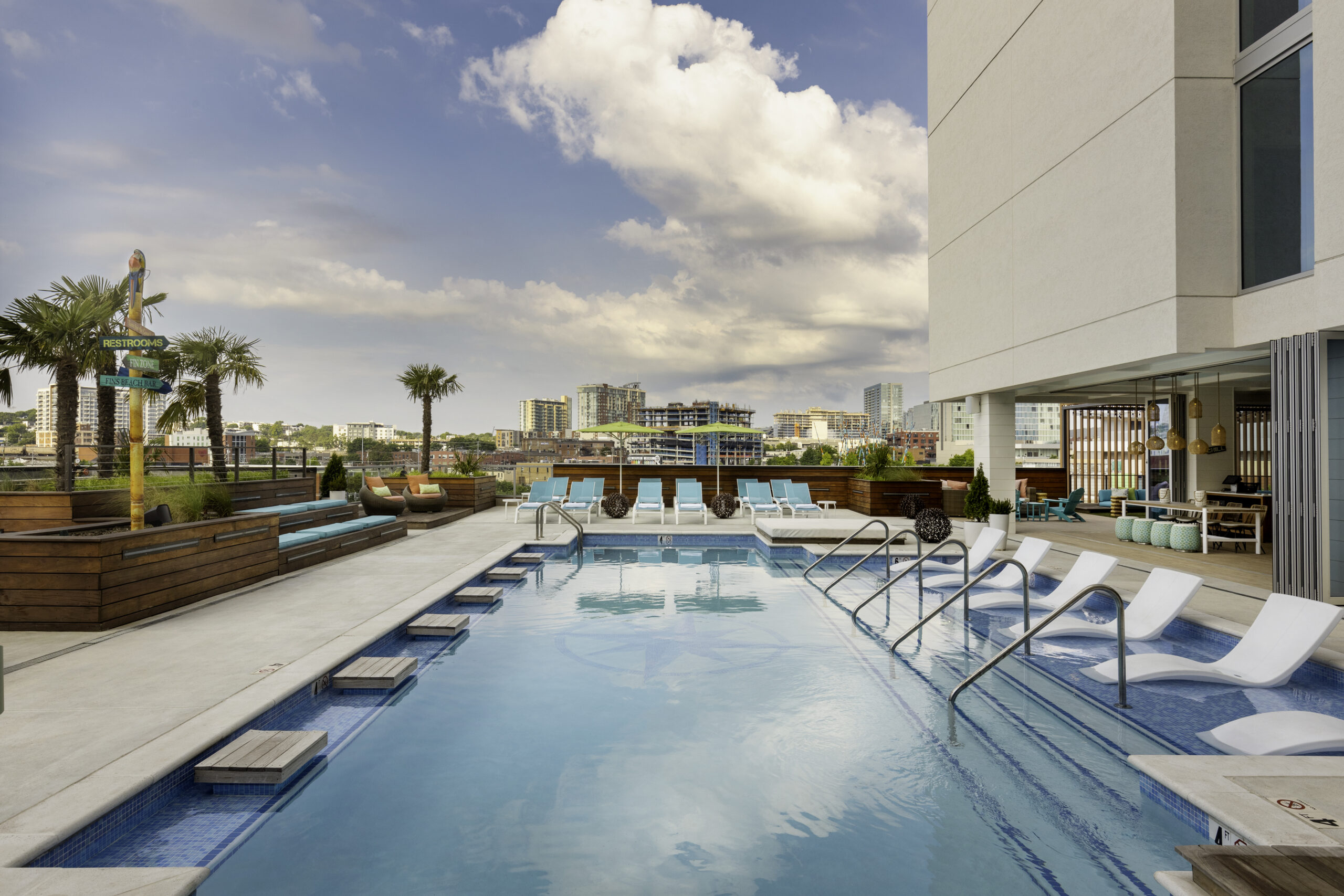 Rooftop pool area with turquoise lounge chairs, palm trees, and a modern building on the right under a blue sky.