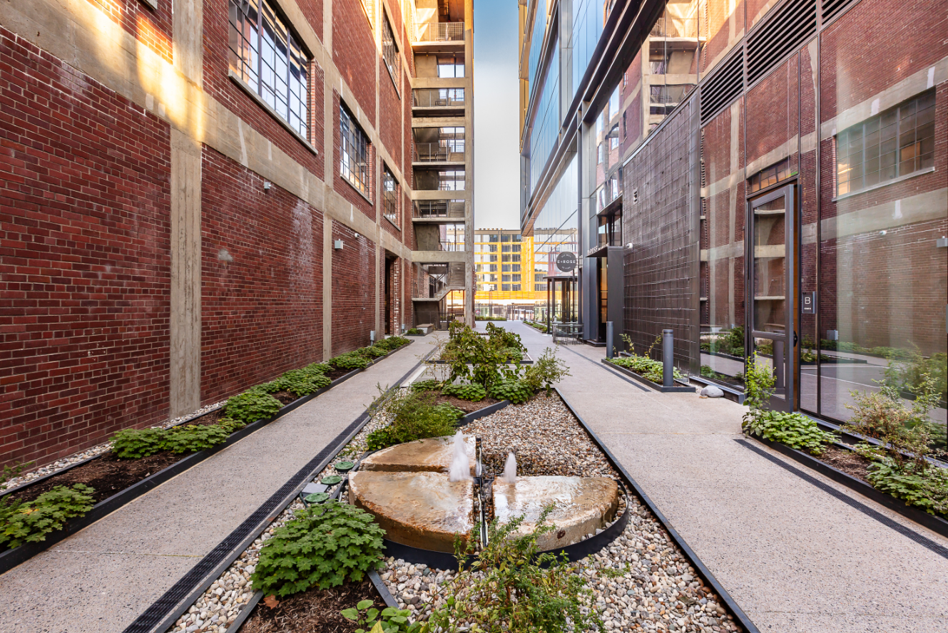 Urban courtyard between brick buildings, featuring a circular stone fountain, gravel beds, and planting along paved walkways.