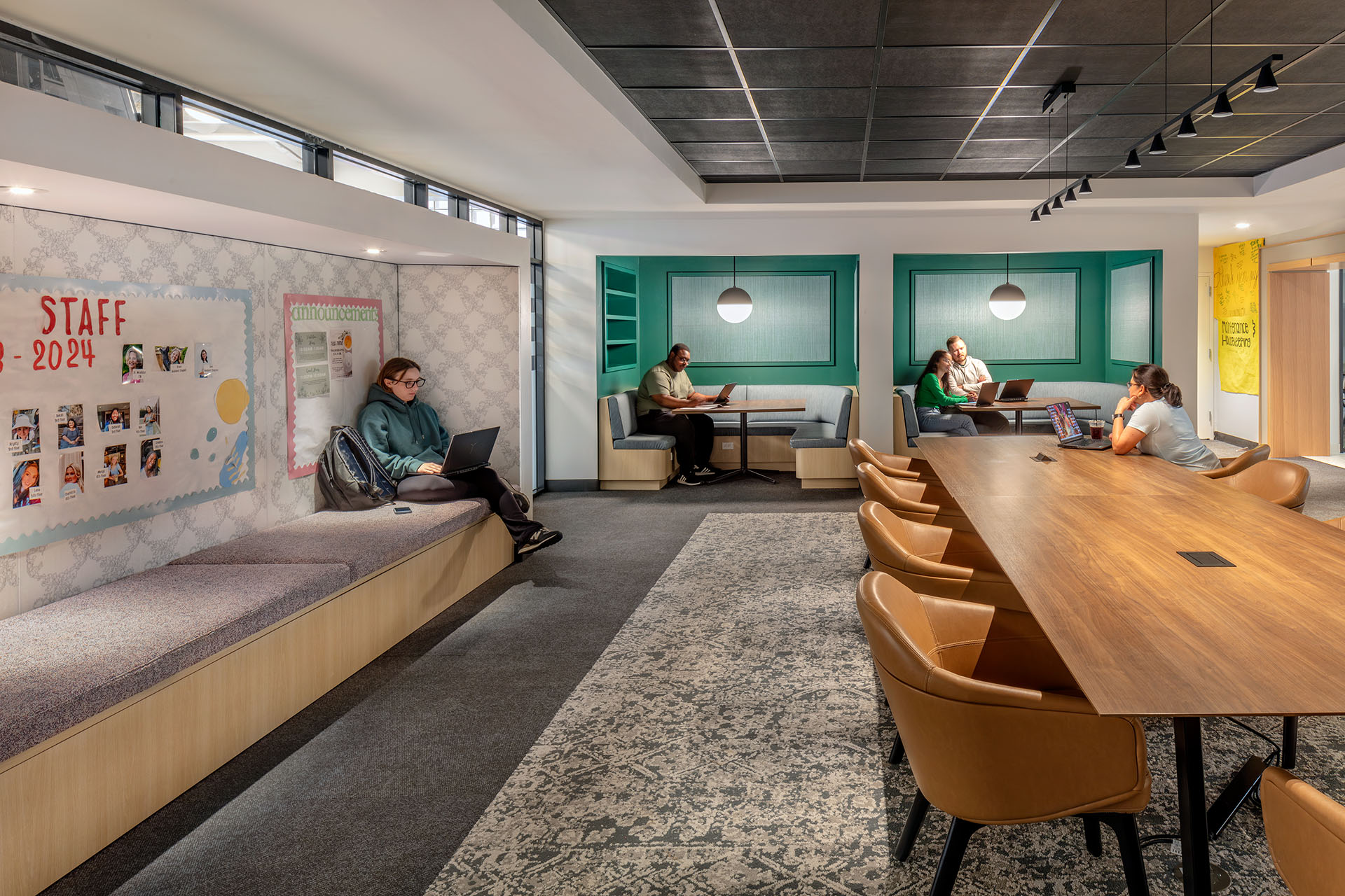A modern study lounge with people using laptops at booth seating and a long wooden table with leather chairs nearby