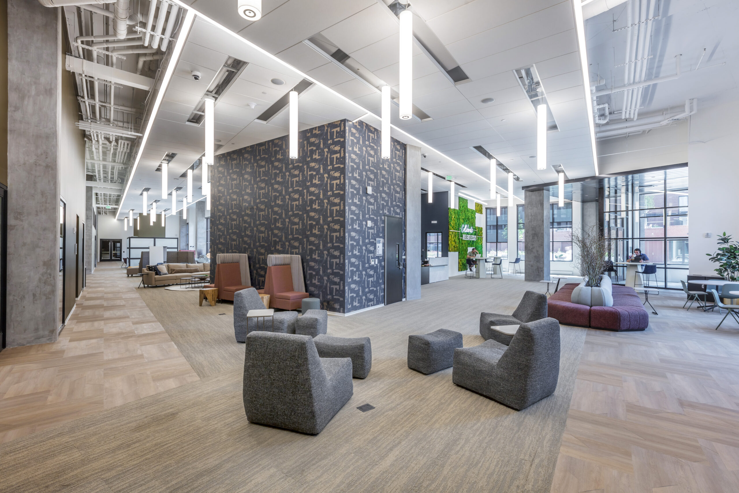 Open, spacious modern lobby with lounge seating, pendant lights, and a patterned dark accent wall; people seated near windows in background.