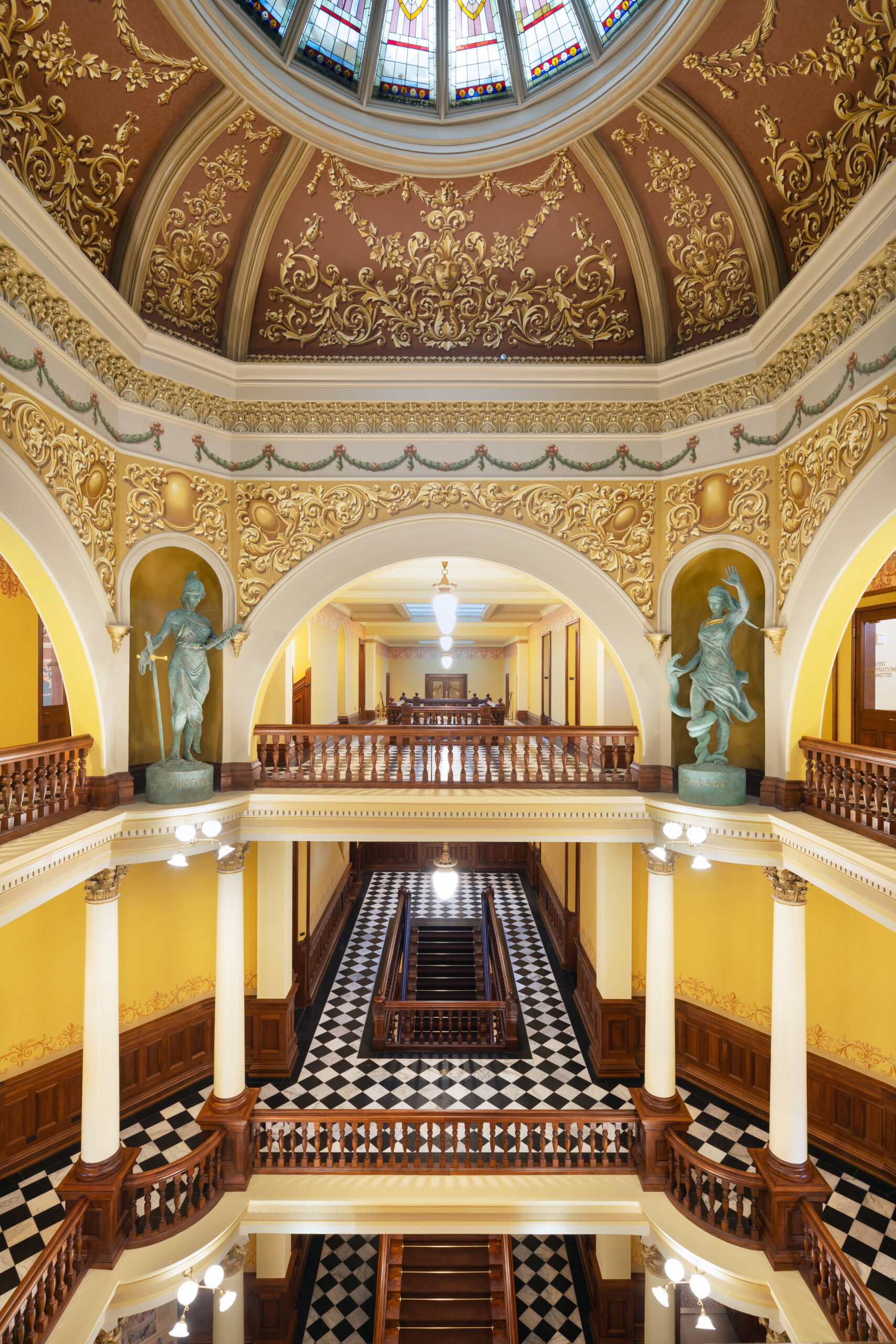 Grand multi-level interior with ornate gold filigree, arches, and a checkered black-and-white floor surrounding a central wooden staircase.