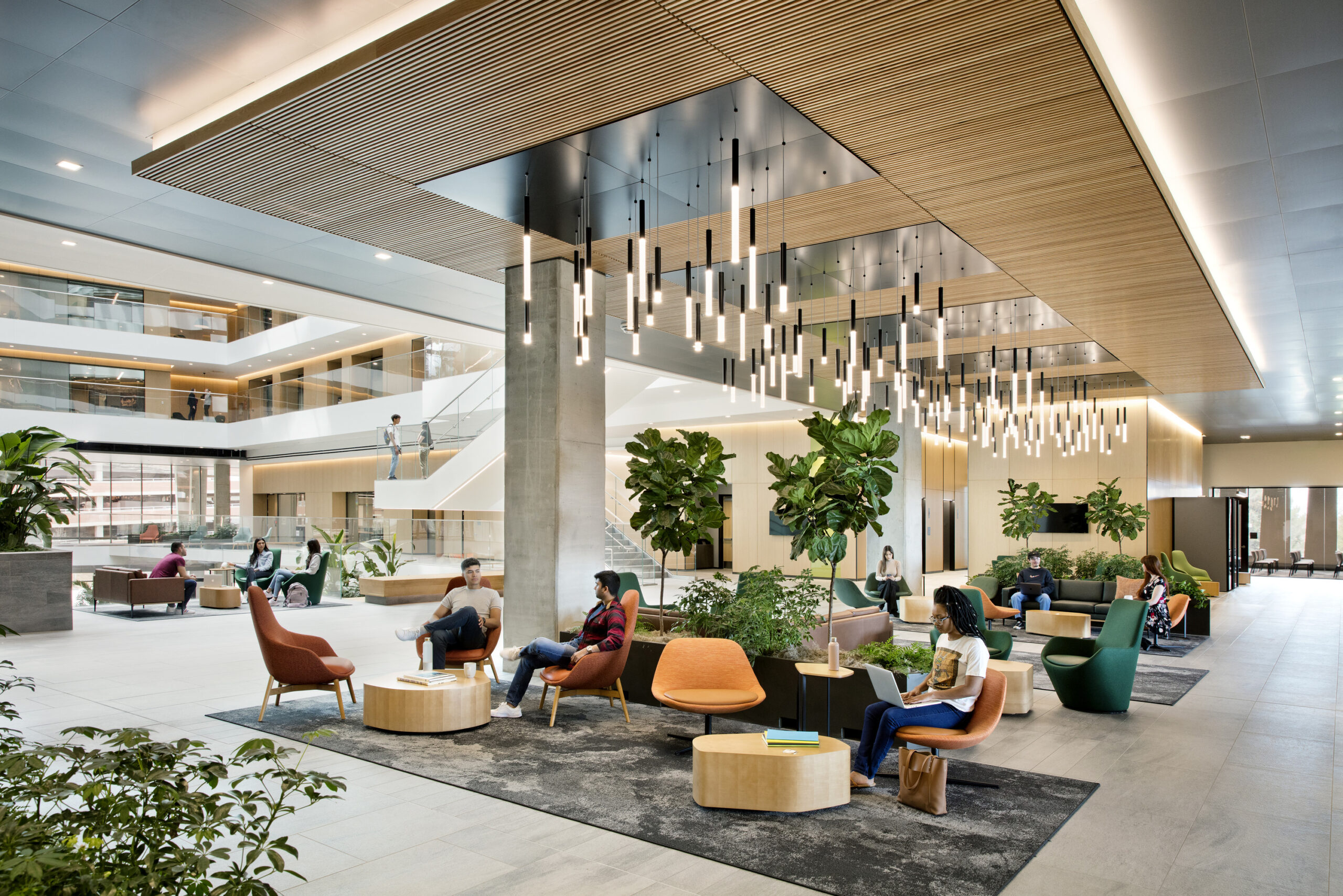 Modern, open office lobby with orange chairs, people seated and conversing near plants and a staircase under a sculptural ceiling lighting installation.