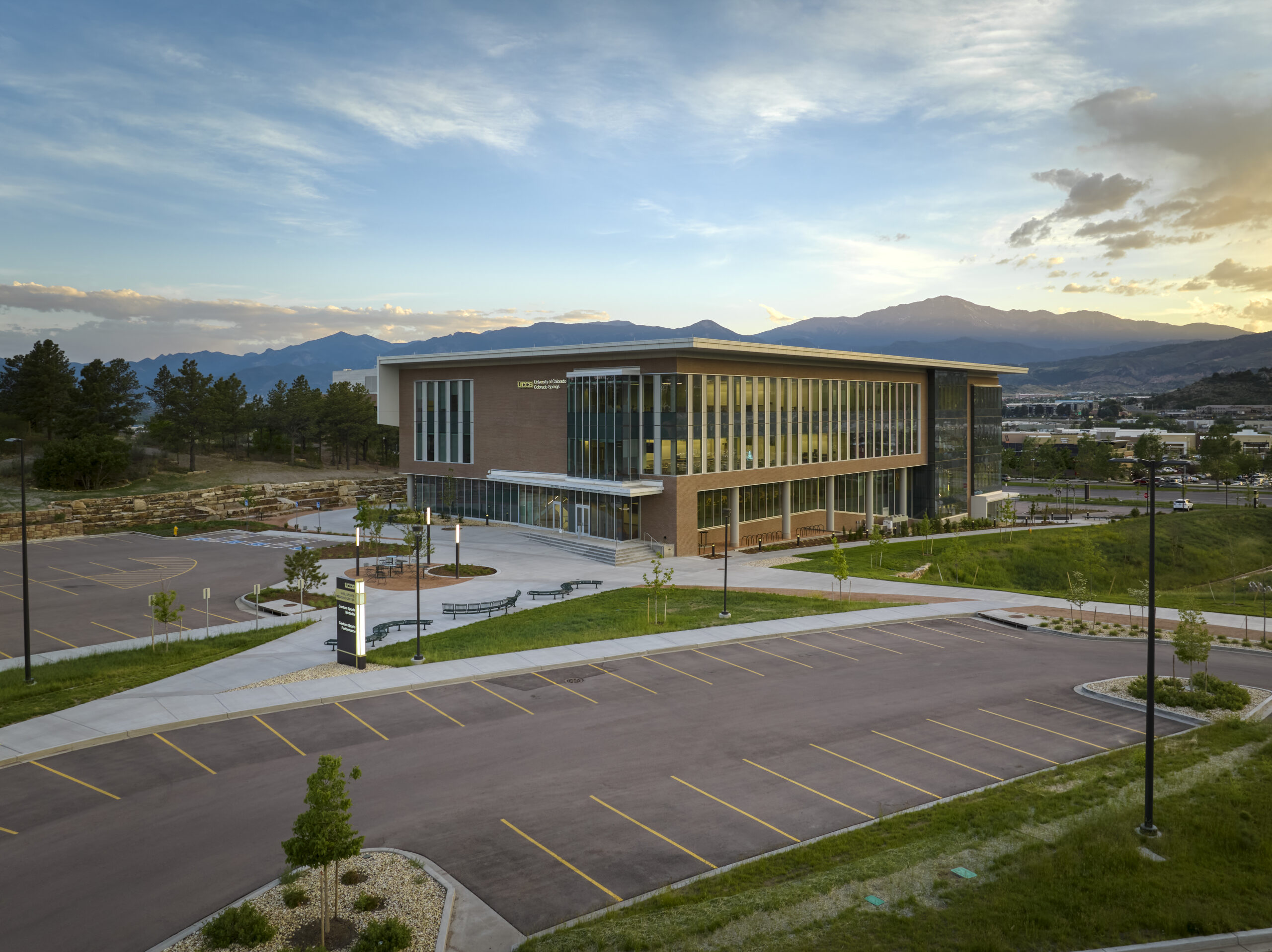 Modern campus building with brick and glass exterior, large windows, and a surrounding parking lot, set against distant mountains and a blue sky.