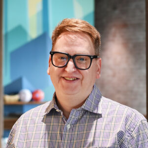 Man with short light brown hair and black glasses smiling at the camera indoors, wearing a checkered shirt.