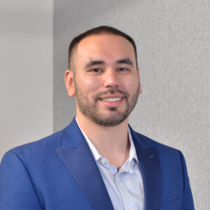 Portrait of a man with short dark hair and a trimmed beard, wearing a blue blazer and a light striped shirt, smiling against a neutral gray background.