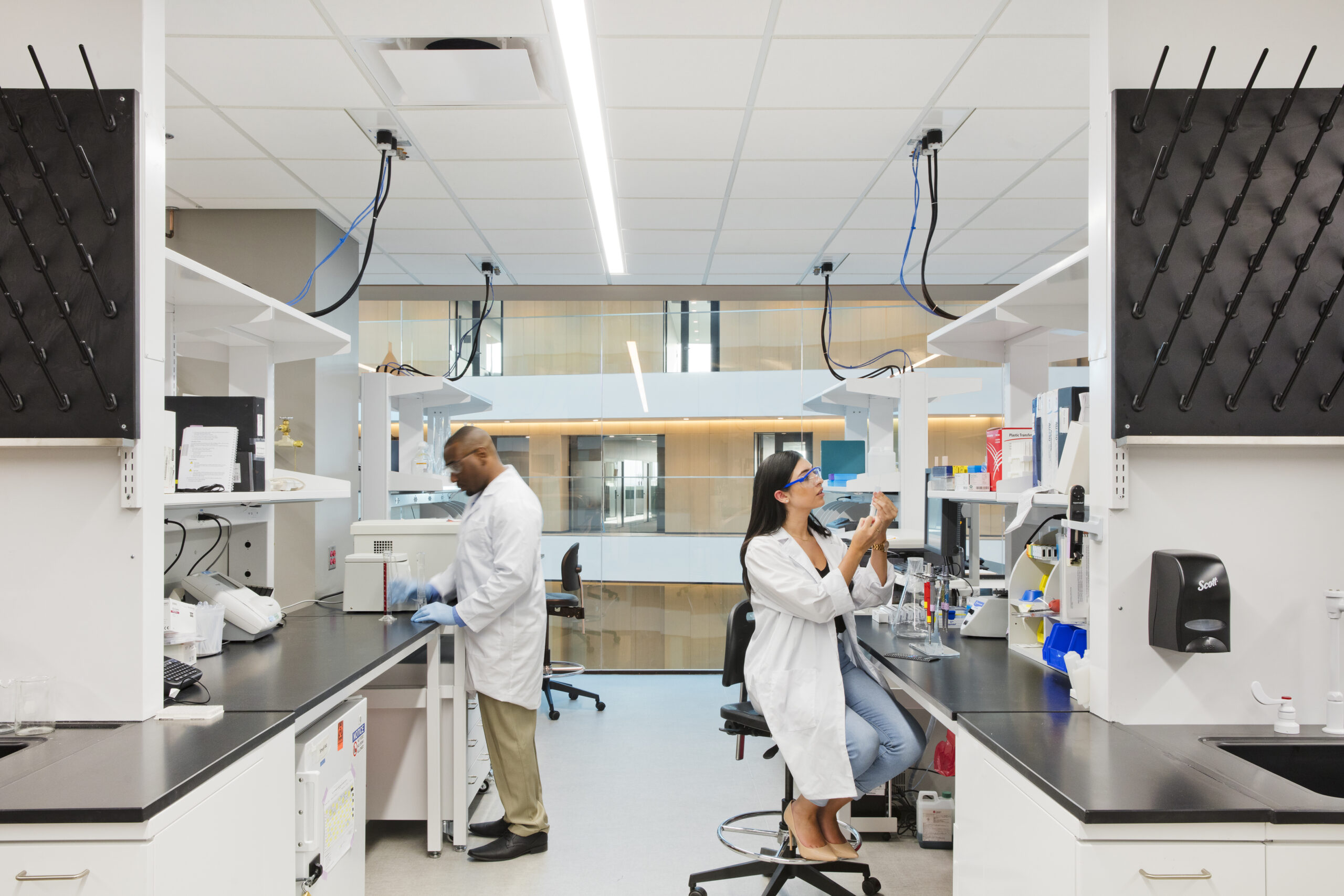Two scientists in white lab coats work at benches in a bright modern laboratory with equipment around them.