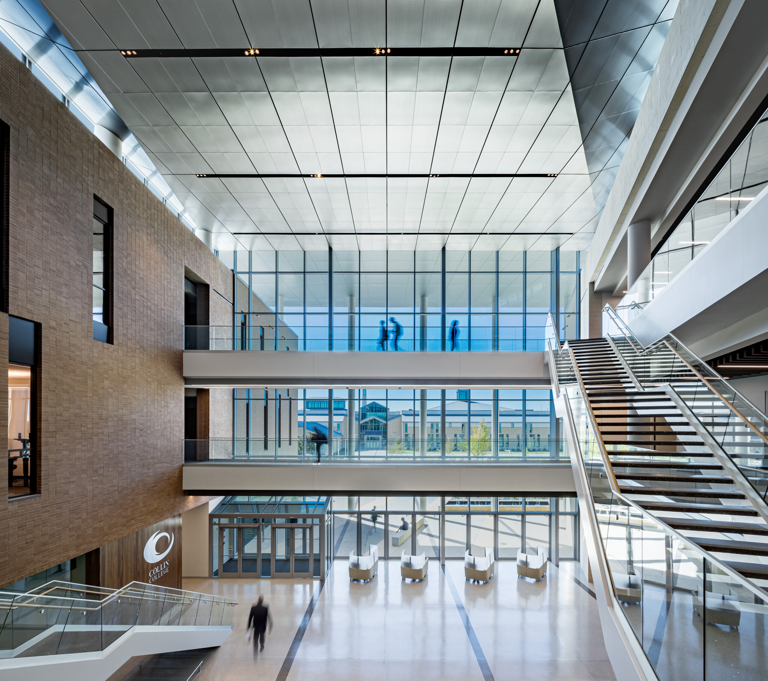 Spacious modern atrium with brick walls, glass railings, and a wide staircase; blurred figures walk across the open space.