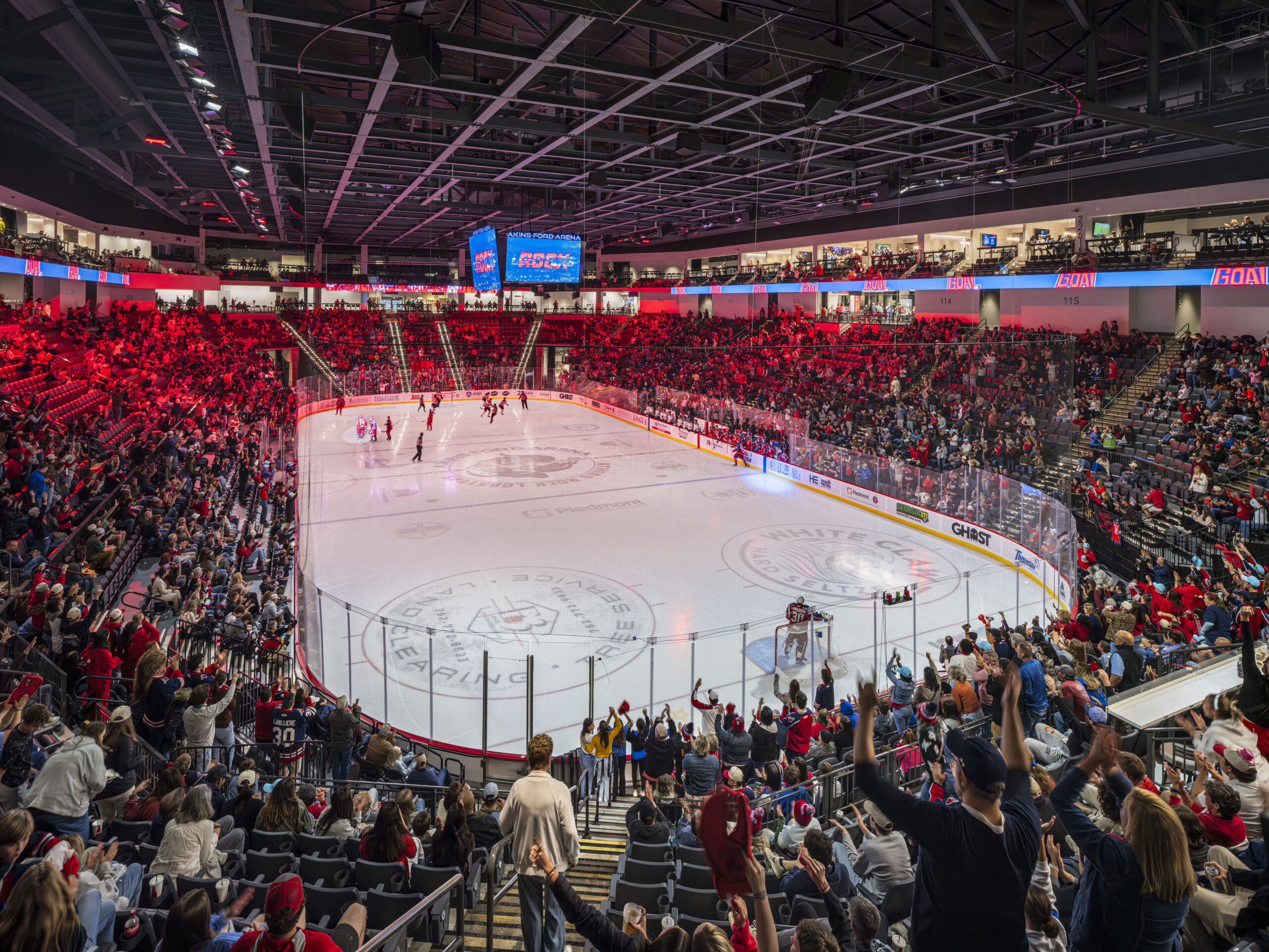 Interior of a lively ice hockey arena filled with fans, teams on the ice, and a large central scoreboard above the rink.