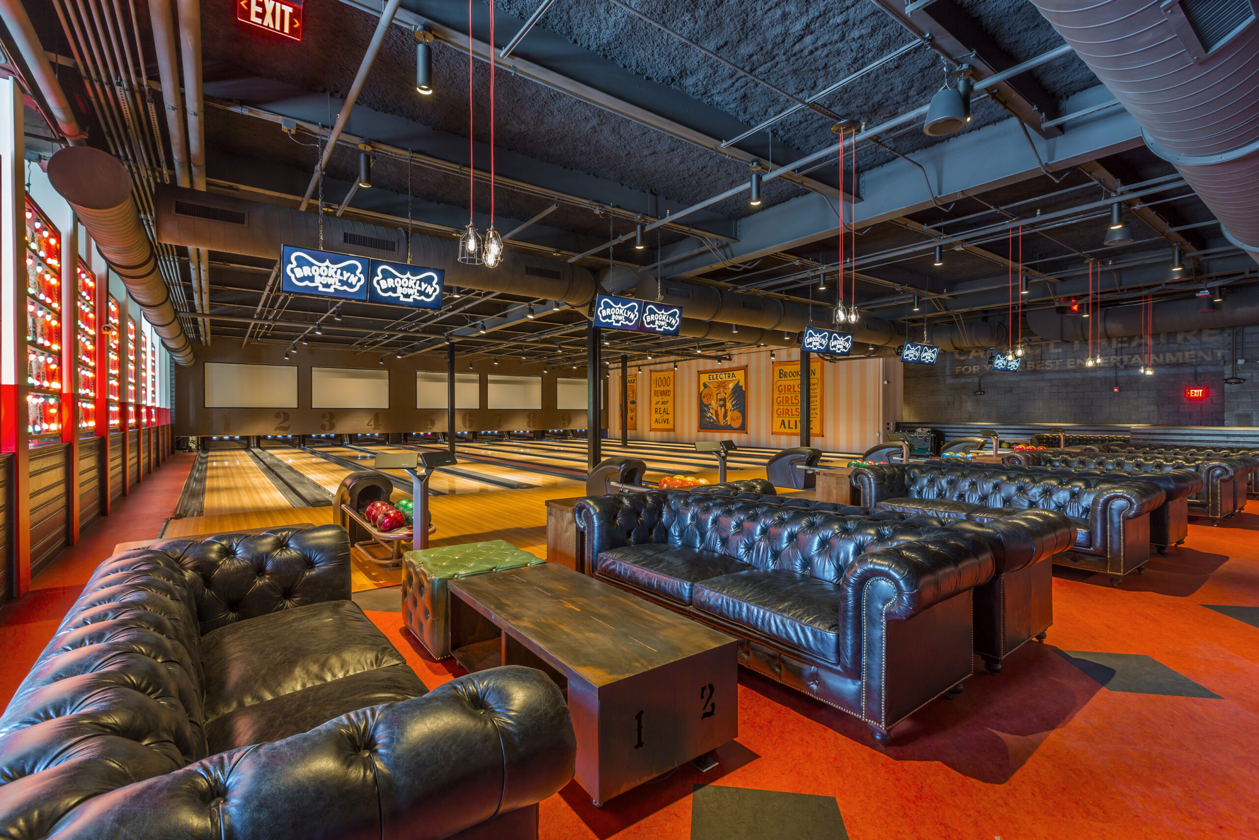 Luxurious bowling lounge with blue tufted leather couches, wooden lanes, and neon Brooklynbowl signs overhead.