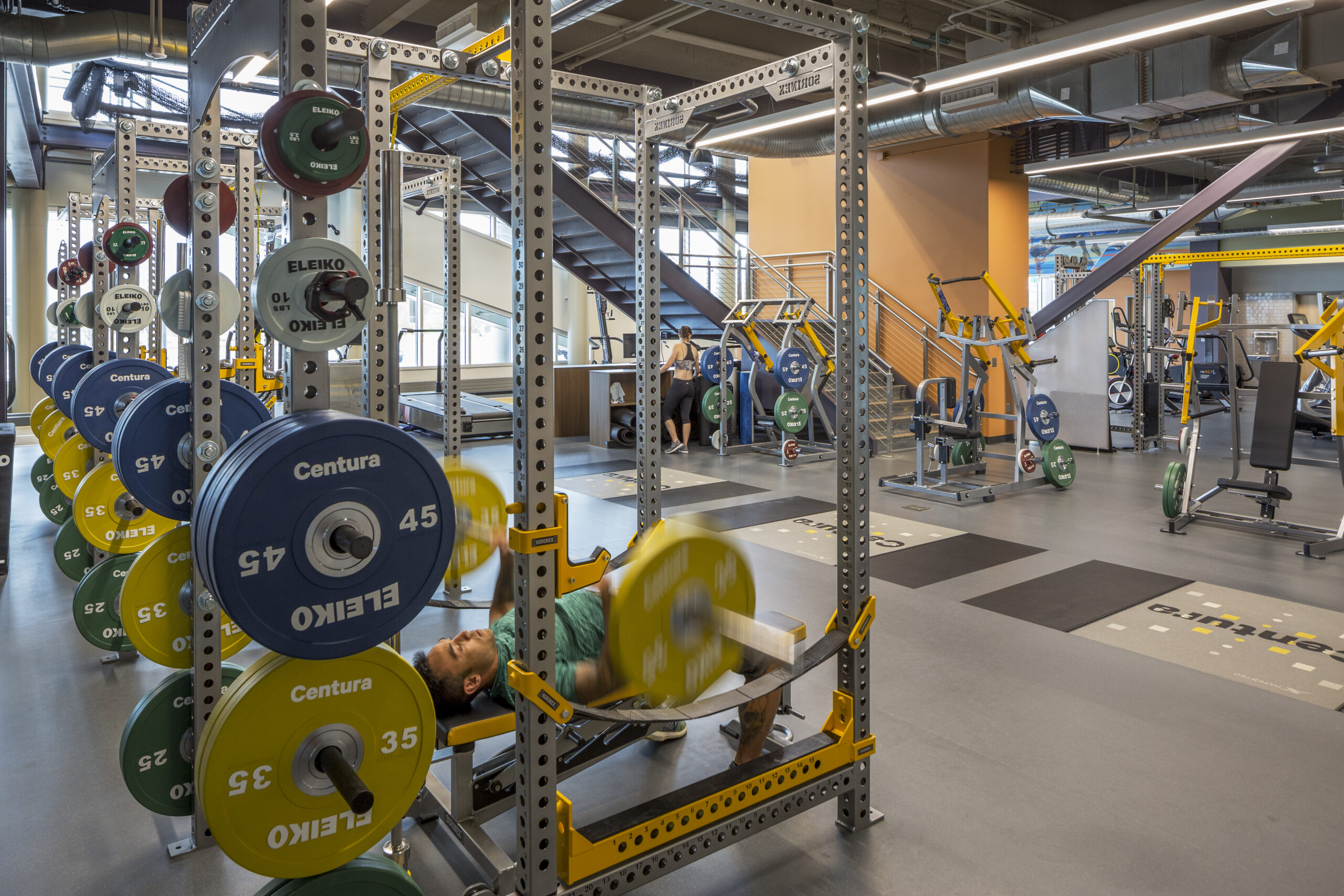 Modern gym with power racks and loaded bumper plates in blue, yellow, green and red; a person bench pressing beneath a squat rack under bright industrial lighting.