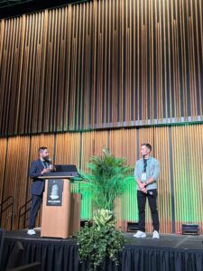 Emory AutoDesk_NexusCon2026 - Aptitude Two men on a stage at a conference; one speaks at a podium with laptop while the other stands nearby, wood slat wall backdrop and green plants behind.