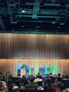 Same Asset One Name - Aptitude Three men speak into microphones on a stage during a conference panel, with an audience at round tables watching.