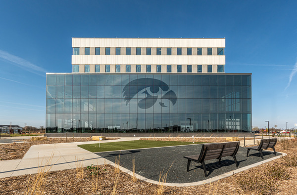 UIHC-121 - Aptitude Modern glass-front building with a large hawk logo on the façade, benches in the foreground on a sunny campus plaza.